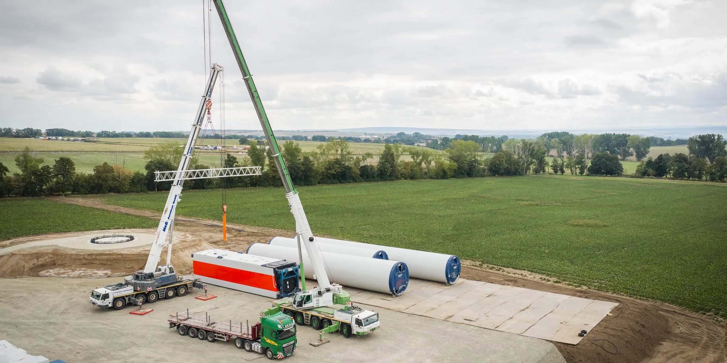 Luftbild einer Windpark-Baustelle: zwei Autokrane heben eine Traverse, daneben liegen Turmsegmente