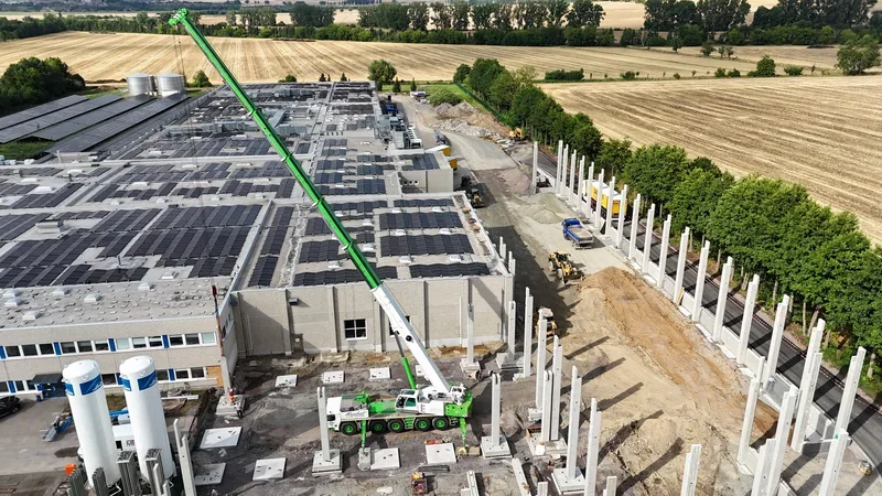 Luftbild einer Baustelle, Autokran stellt Betonstützen neben einer Halle