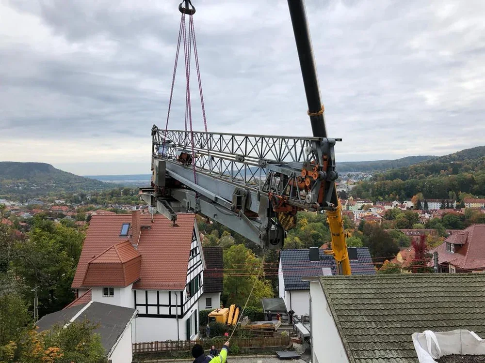 Autokran hebt ein Auslegerteil über den Dächern eines Ortes, dahinter Berge