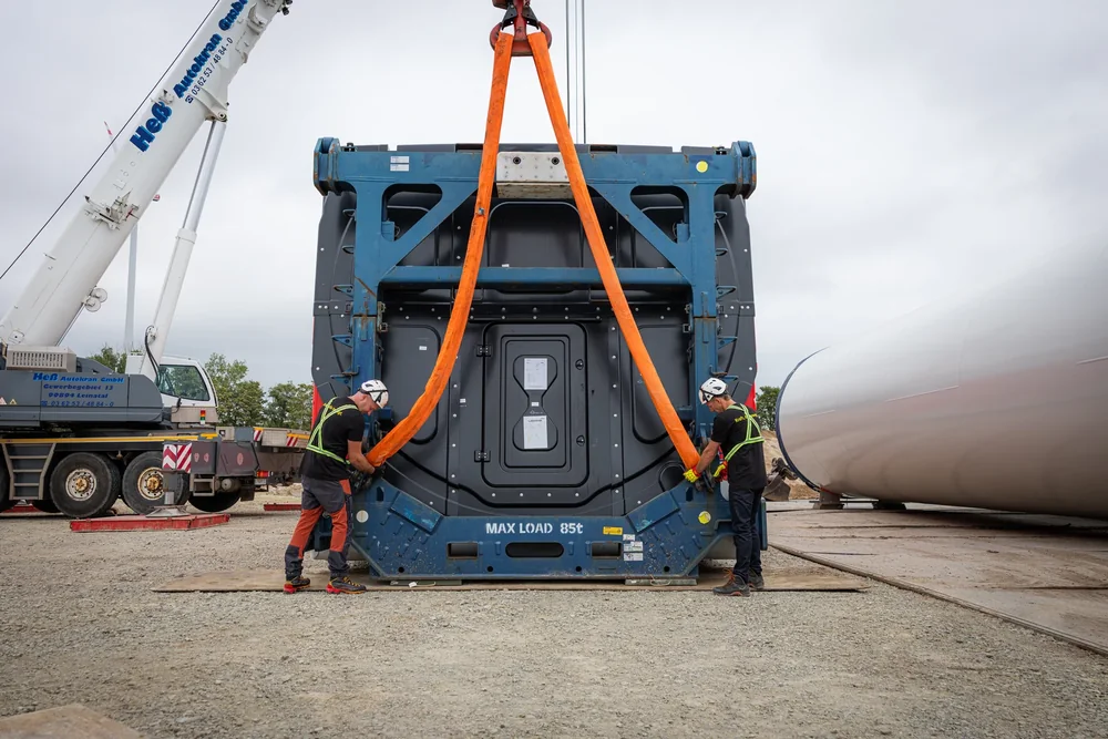 Zwei Anschläger schlagen ein großes Windkraft-Bauteil in seinem Transportgestell mit orangen Rundschlingen am Kranhaken an