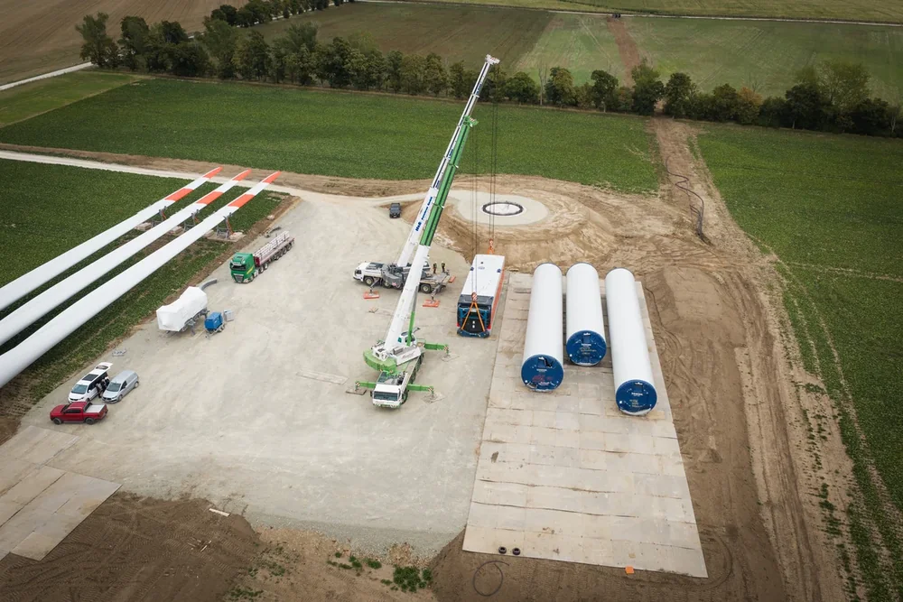 Luftbild einer Windpark-Baustelle: Autokran neben Rotorblättern und Turmsegmenten auf der Kranstellfläche